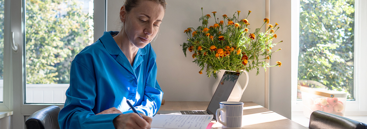 Woman in blue shirt journaling at a sunlit table with a laptop, coffee mug, and vase of marigolds nearby.