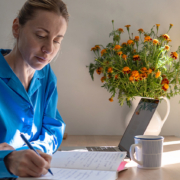 Woman in blue shirt journaling at a sunlit table with a laptop, coffee mug, and vase of marigolds nearby.