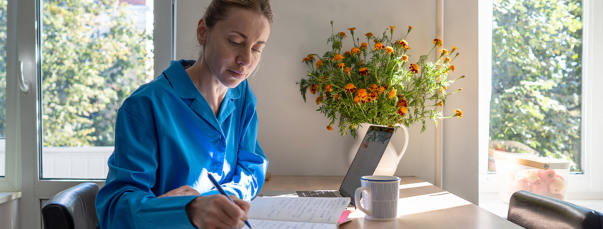 Woman in blue shirt journaling at a sunlit table with a laptop, coffee mug, and vase of marigolds nearby.