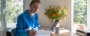 Woman in blue shirt journaling at a sunlit table with a laptop, coffee mug, and vase of marigolds nearby.